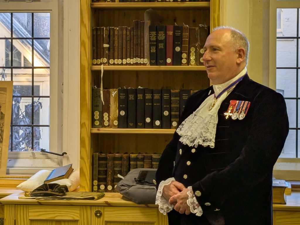Photograph of John May in his uniform as High Sheriff of Oxfordshire. He is stood in front of a wooden bookshelf in the new Gibbs Library.